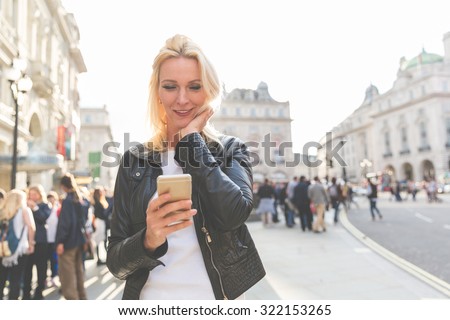 Adult woman looking at smart phone in London at sunset. She is a blonde woman on her early forties, she looks candid and spontaneous. Backlight shot with blurred people on background.