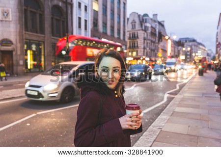 Young woman in London standing by a busy road at night. She is on her mid twenties, holding a cup of coffee or tea and looking at camera. Her face is illuminated by traffic light.