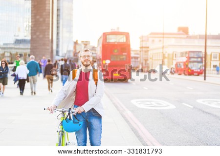 Hipster man walking on London bridge and holding his fixed gear bike. He is walking on the sidewalk, and there are some people and red buses on background. Lifestyle and transportation concepts.
