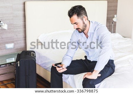 Businessman looking at smart phone in his hotel room. He is sitting on the bed, wearing black trousers and a light blue shirt. Grave expression, business and work issues concepts.