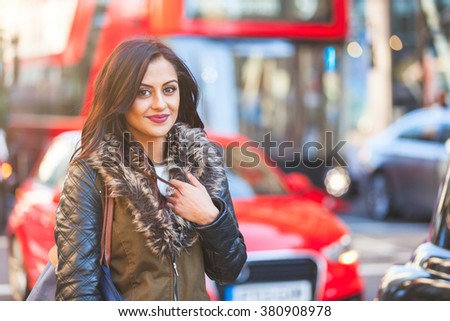 Indian girl portrait in London. She is standing by a busy road with blurred traffic on background. There are cars and red buses. She is smiling and looking at camera. Travel and lifestyle concepts