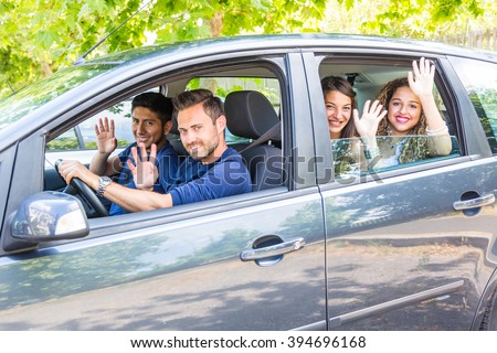 Group of people in the car. They are a multicultural group of friends leaving for a trip. There are two men sitting on the front and two women on the back. They are smiling and waving their hands.