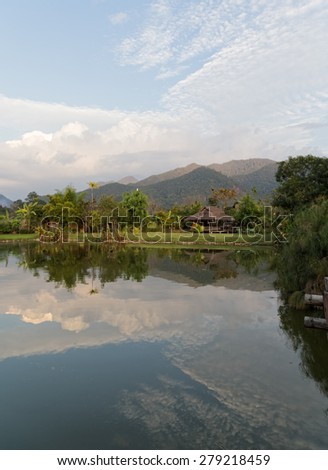 Reflection of a bamboo hut and mountain and trees on the water of a pond in rural Thailand.
