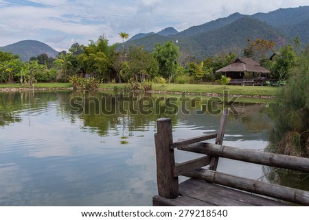 Reflection of a bamboo hut and mountain and trees on the water of a pond in rural Thailand. Serene with wooden dock in the foreground.
