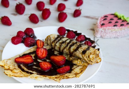 Pancakes with strawberries and chocolate on a white plate and a cake in the background on a table