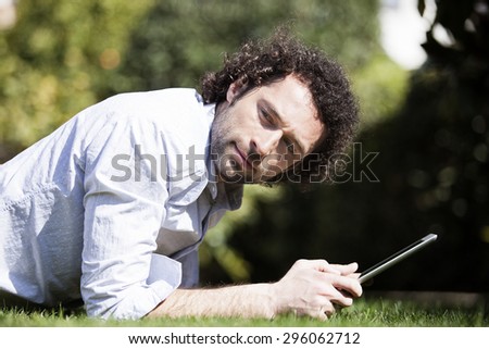 Handsome young smiling man lying on grass at park and using digital tablet