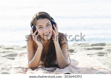 Young attractive woman listening to music on sand beach, relaxing on a towel while on vacation and smiling on a sunny day.