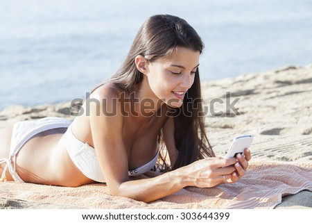 Portrait of an attractive girl laying down relaxing on holiday, using a smartphone device on vacation laying down on a beach towel on the sand. People travel technology.