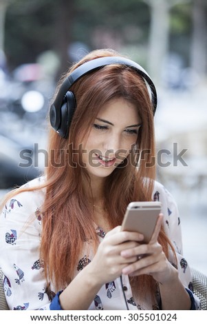 Gorgeous female enjoying recreation time while listen some music with her mobile phone on terrace bar, smiling successful young woman  looking at the phone screen