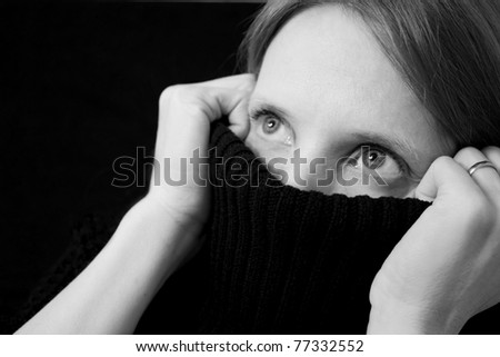 Black and white image of a young, pretty woman partially covering her mouth and nose with her sweater, looking up and out of the frame towards black background with copy space
