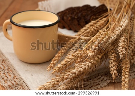 Armful of ears, fresh bread and a metal mug of milk on a wooden surface covered with a linen napkin