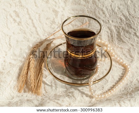 A glass cup of black tea, a pearl necklace and several mature ears of wheat on a cream lace surface