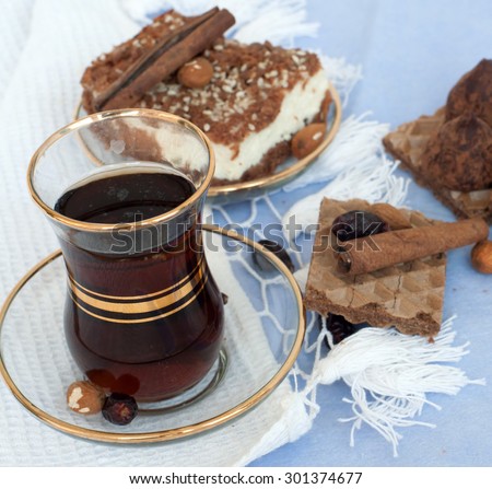 A glass of black tea, a piece of a chocolate pie on a glass saucer, waffles, truffles and cinnamon sticks on a white and blue table cloth