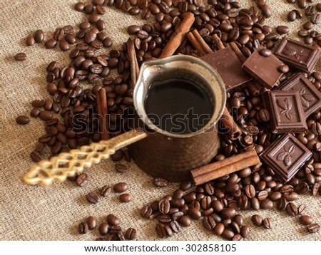 A coffee pot, roasted coffee beans, cinnamon sticks and pieces of chocolate on a sackcloth, close up