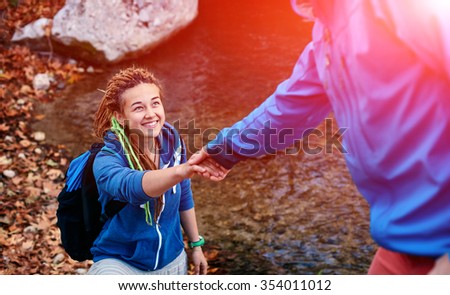 Helping hand - hiker woman getting help on hike smiling happy overcoming obstacle. Tourist backpackers walking in autumn forest. Young couple traveling.