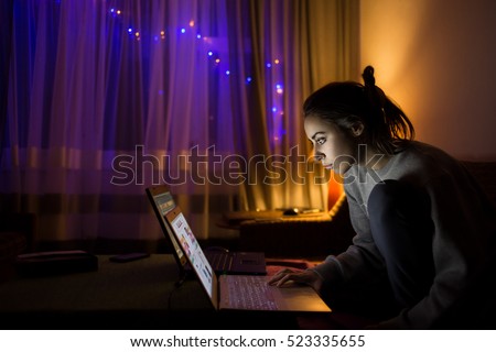 woman watching laptop at winter evening in dark room with window as background. illuminated garland in the window