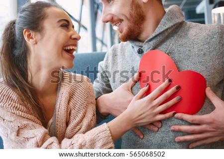 man and woman smiling and holding a large paper heart. Two people in cafe communicate, laughing and enjoying the time spending with each other. Couple in love on a date.  Valentines Day concept