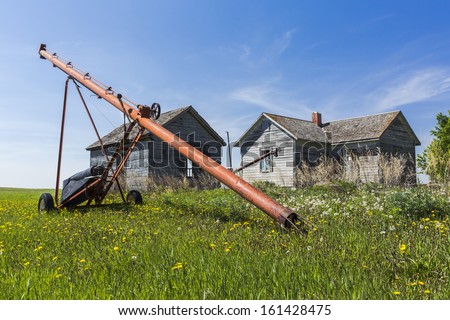 An old orange and red grain auger on a farm yard by some old rustic buildings