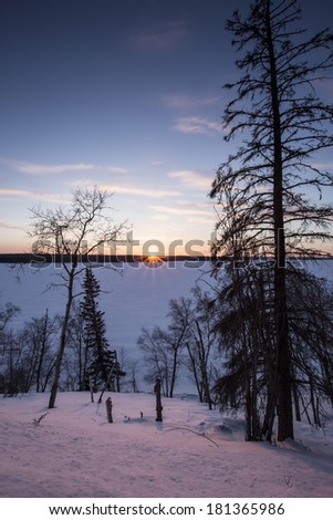 The sun sets over a frozen snow covered lake with trees in silhouette in the foreground.
