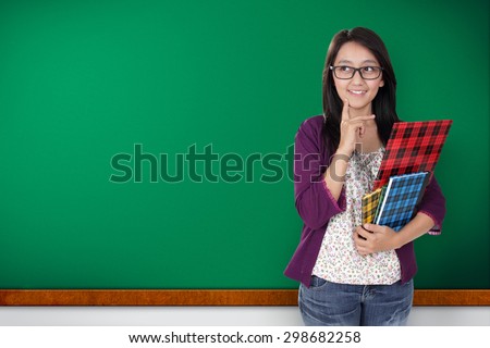 Friendly Asian teacher holding some books, looking to her side, smiling and thinking of a plan, over green chalk board background for copy space