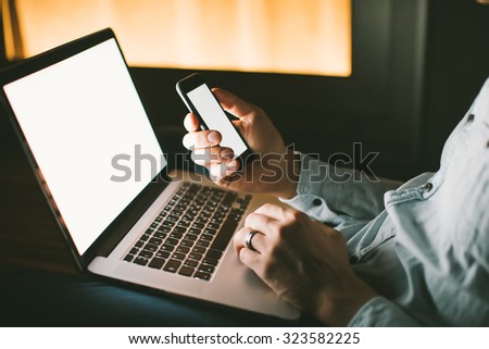 Close up of man's hands holding smartphone with blank screen and using laptop with blank screen