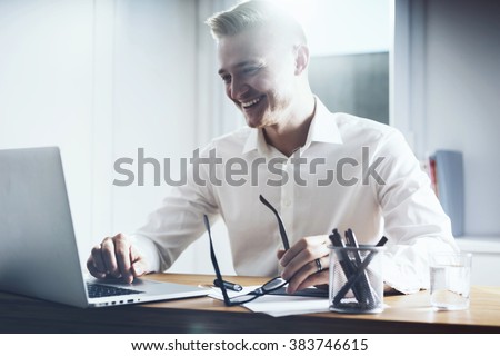 Happy businessman working at his office with laptop. Young smiling man sitting at his workplace with notebook computer and holding glasses in hand