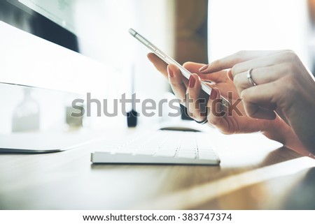 Close-up of female hands using modern smart phone while working at office with computer, businesswoman typing text message on her cellphone, flare light