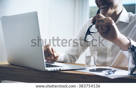 Smiling businessman sitting at his office and using laptop, holding glasses in hand, smart phone on desk, close up image of happy smiling man working on computer