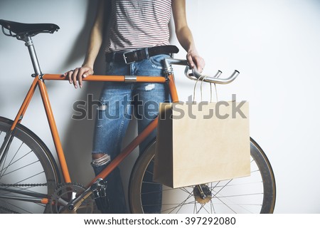 Closeup of woman wearing blue jeans and standing with vintage orange bike and blank paper package, mock-up of craft shopping bag with handles