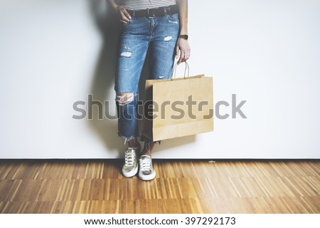 Close-up of hipster girl wearing blue jeans and holding blank craft paper package, mock-up of template brown shopping bag with handles