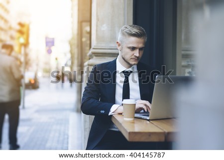 Handsome businessman wearing suit and using modern laptop outdoors, successful manager working in cafe during break and searching information in internet on his notebook computer