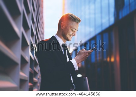 Happy smiling businessman wearing black suit and using modern smartphone near office at early morning, successful employer to make a deal while standing near skyscraper office at night, flare light