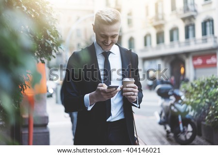 Happy smiling businessman going to the work with coffee cup and using modern smartphone outdoors, successful employer using cellphone while on break outside at sunny day near his office
