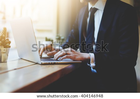Male hands using laptop in modern coffee shop or loft, professional businessman in black suit working on new project with notebook computer while sitting at his office, flare light, blurred background