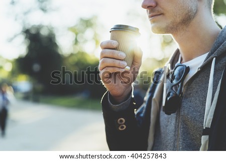 Close-up of young man holding coffee to take away at early morning in sunny park, sunlight, blurred background