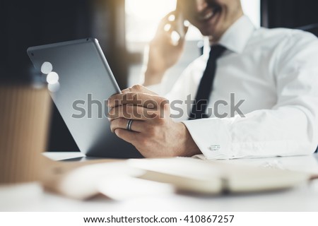 Close-up of happy smiling businessman or employer working on digital tablet and talking on his smartphone in modern office interior, flare light