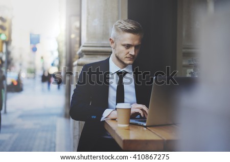 Young professional businessman wearing suit and using modern laptop outdoors, successful manager working in cafe during break and searching information in internet on his portable computer