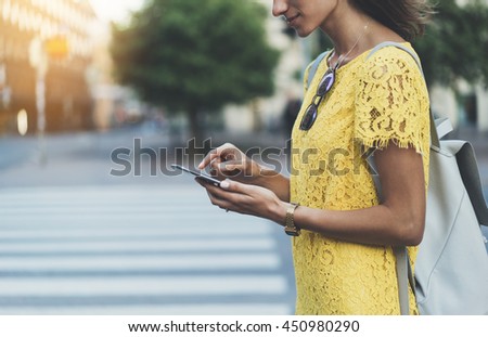 Happy smiling young girl using modern smart phone while standing at crosswalk, hipster girl writing text message on internet via cellphone, attractive woman wearing yellow dress, walking in the city
