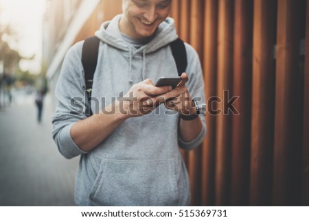 Happy smiling hipster guy chatting with friends at social networks, young cheerful man using modern smartphone device while walking at city street, urban lifestyle, flare light