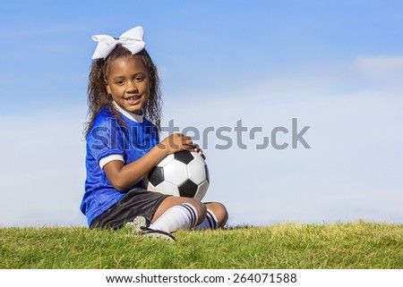 Cute, young african american girl soccer player holding a ball sitting on a grass field