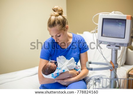 Young mother holding her Premature newborn baby who is being treated in the hospital. With love and tenderness she holds her baby close while sitting on a hospital bed next to a vital signs monitor