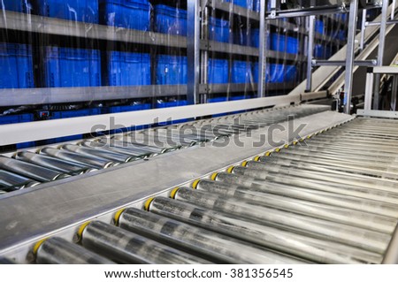 Close up shot of two roller conveyors in an automated warehouse with a ramp and piles of blue plastic boxes in the background. Photo taken in a big warehouse in Germany. Focus on the conveyor rolls.