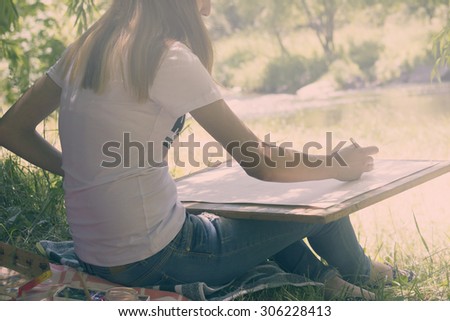 Young woman draws on an easel against the river and the forest background