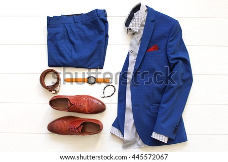 Flat lay set of classic men`s clothes such as blue suit, brown shoes, belt, watches and bracelet on white wooden background. Top view.