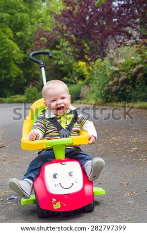 Baby boy is playing in the garden on a little car. He is smiling.
