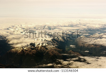 Mountain view from the top through the clouds.   The mountain peaks covered with snow. View from airplane. sunlight tint