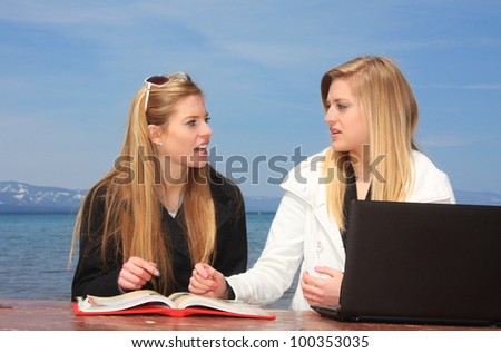 Two teen aged girl students sitting at a park bench studying with a book and computer.