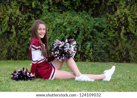 Pretty young cheerleader wearing a maroon colored uniform and pom-poms posing outdoors on lawn.