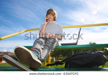 Young teen aged boy talking on cell phone, sitting on bleachers with a laptop beside him.