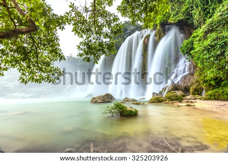 Played with the moss rock waterfall Ban Gioc with lakeside tree foreground, smooth flow of water, creating a waterfall down the white silk sheet side high mountains, sunny yellow iridescent great lake